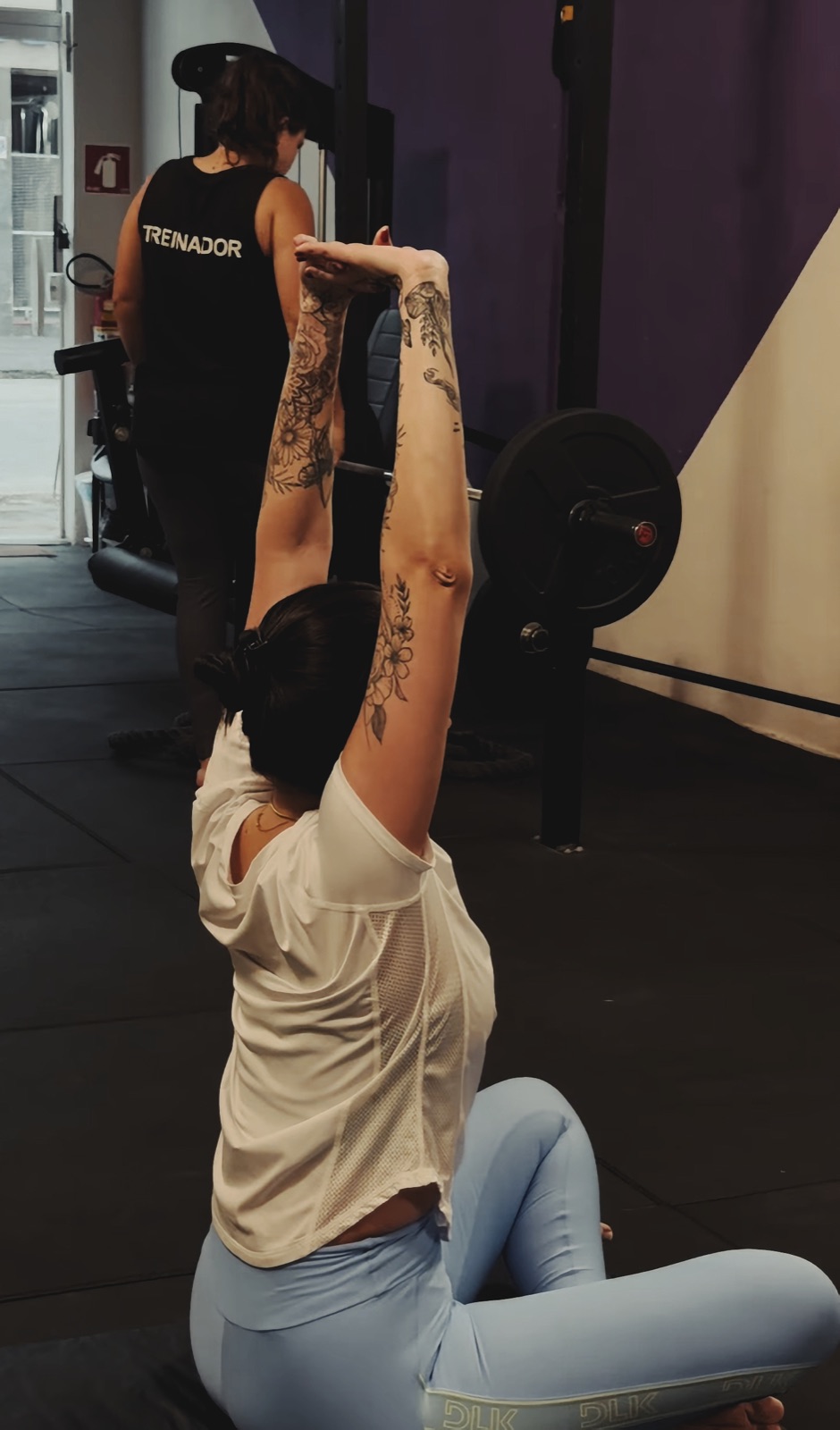 Aluna treinando com barra, acompanhada por um treinador